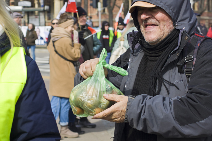 Пожилой мужчина в темной одежде с пакетом овощей в руках.