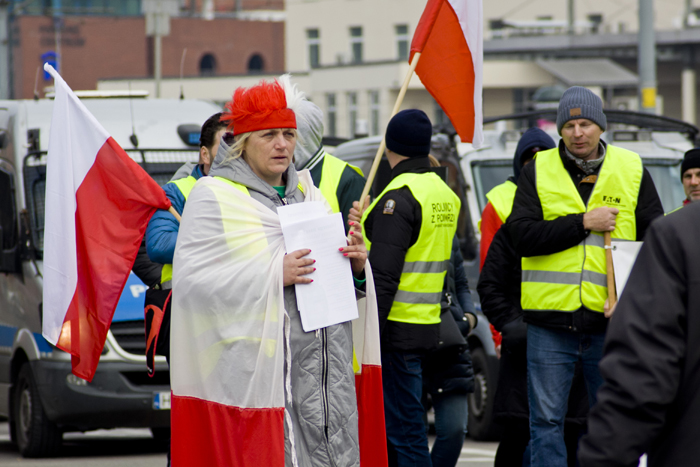 Учасниця протесту фермерів з державним прапором Польщі на плечах на фоні протестувальників в жовтих жилетах.