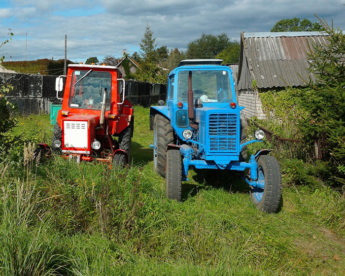 Два небольших трактора красного и голубого цвета стоят на зеленой траве около небольшого дома.