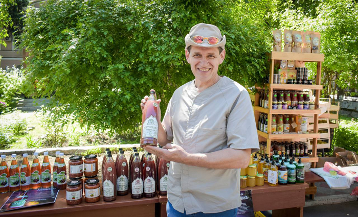 Предприниматель Иван Дегтяр стоит возле стола и стеллажа, где выставлена продукция его предприятия в бутылках и банках.