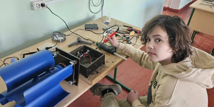Школьник из города Каменец-Подольский Артем Ясинский сидит за столом с разными приборами и инструментами и мастерит вертикальный ветрогенератор.